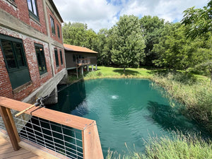 A high-angle view shows a vibrant, teal-colored pond situated next to a multi-story red brick building with green-trimmed windows. A wooden deck with a fishing rod is in the foreground. In the background, there is a brown house on stilts, green grass, and dense green deciduous trees under a cloudy sky.
