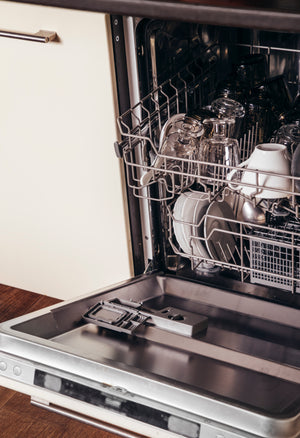 An open dishwasher reveals a rack filled with clean dishes. The top rack contains several upside-down clear and dark-tinted glasses alongside a white ceramic mug. The bottom section shows white ceramic plates standing vertically and a grey plastic cutlery basket. The dishwasher door is horizontal, showing the detergent dispenser.