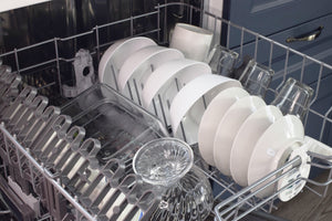 A gray metal upper dishwasher rack is filled with various clean white ceramic bowls, small plates, and clear glasses. A large, rectangular glass container sits on the left side. The dishwasher is partially pulled out, revealing a dark blue kitchen cabinet with a bronze handle in the background.