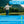 An elderly man wearing a tan hat and shirt stands by a pond, tossing a small square packet into the water. The background shows green trees and mountains. The image includes text instructions for 