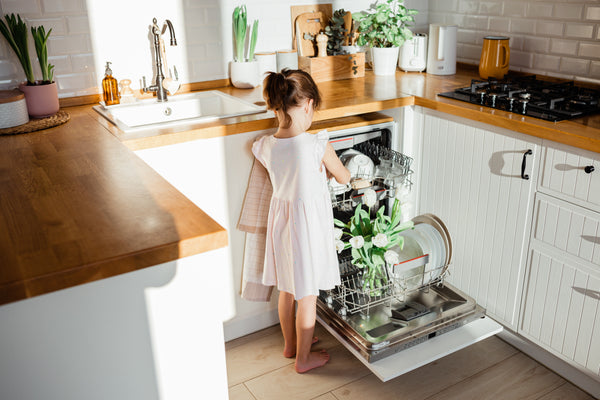 A young girl with brown hair in a ponytail wears a light pink dress while standing in a bright kitchen. She is placing a bouquet of white tulips into the bottom rack of an open dishwasher. The kitchen features white subway tiles, wooden countertops, a white sink, and various potted plants.