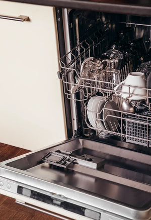 An open kitchen dishwasher loaded with clean glass cups, a white ceramic mug, and ceramic plates, showing the interior racks and an open detergent dispenser.